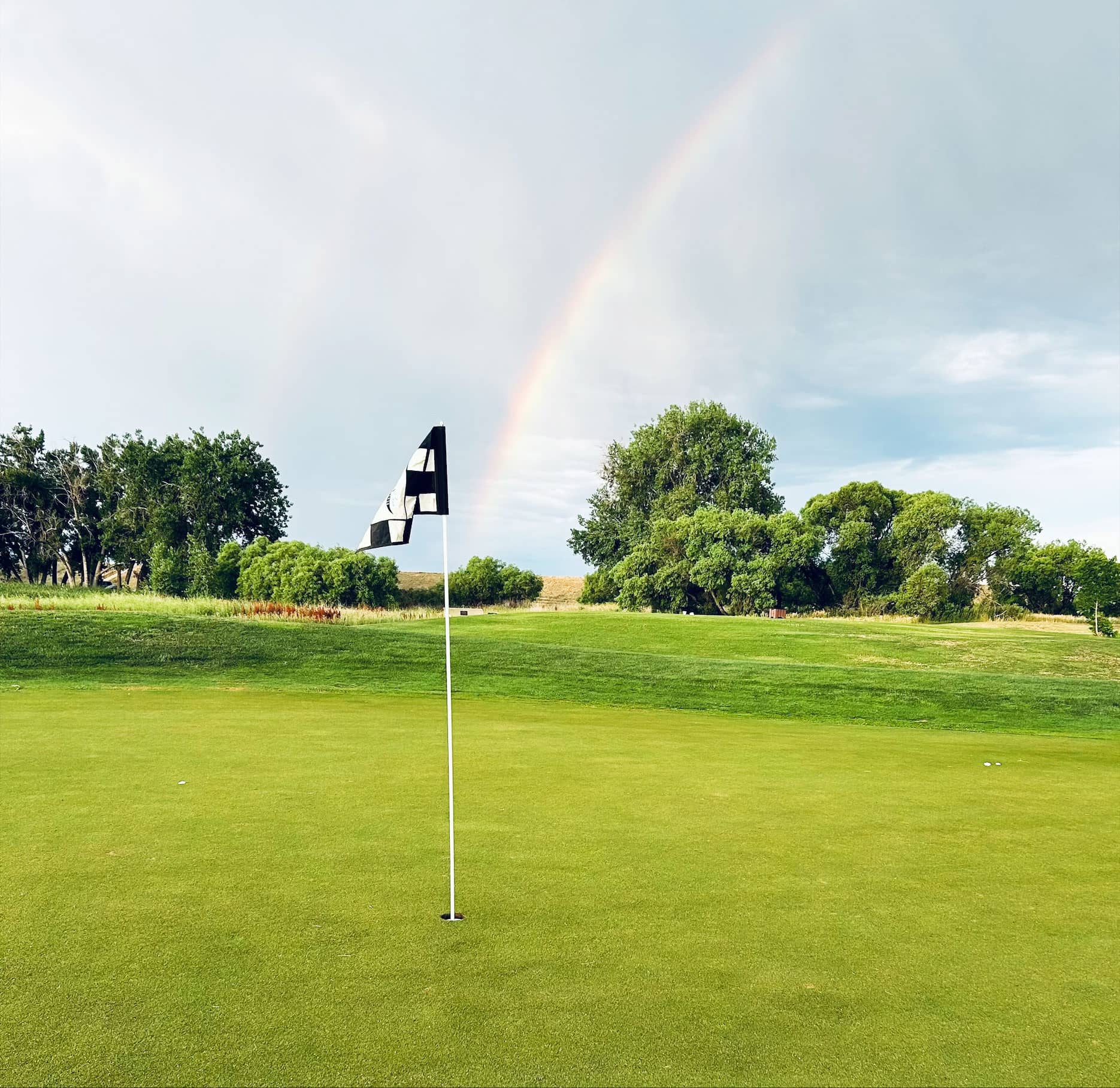 Golf course with a flag and a rainbow in the background, surrounded by lush greenery.
