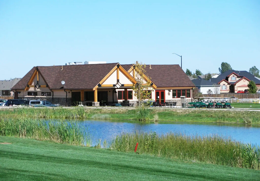 A scenic view of a clubhouse by a pond, surrounded by grass and residential buildings in the background.