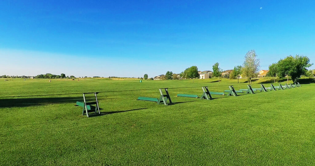 A wide grassy field with green benches lined up, under a clear blue sky with a few trees and houses in the background.