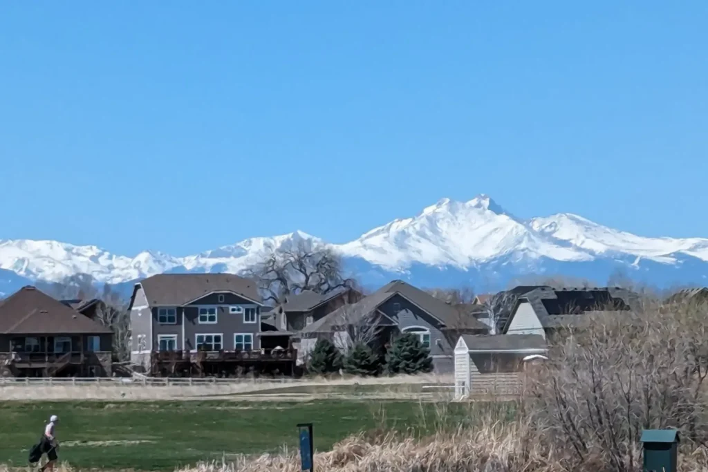 Snow-capped mountains in the background with residential houses in the foreground under a clear blue sky.
