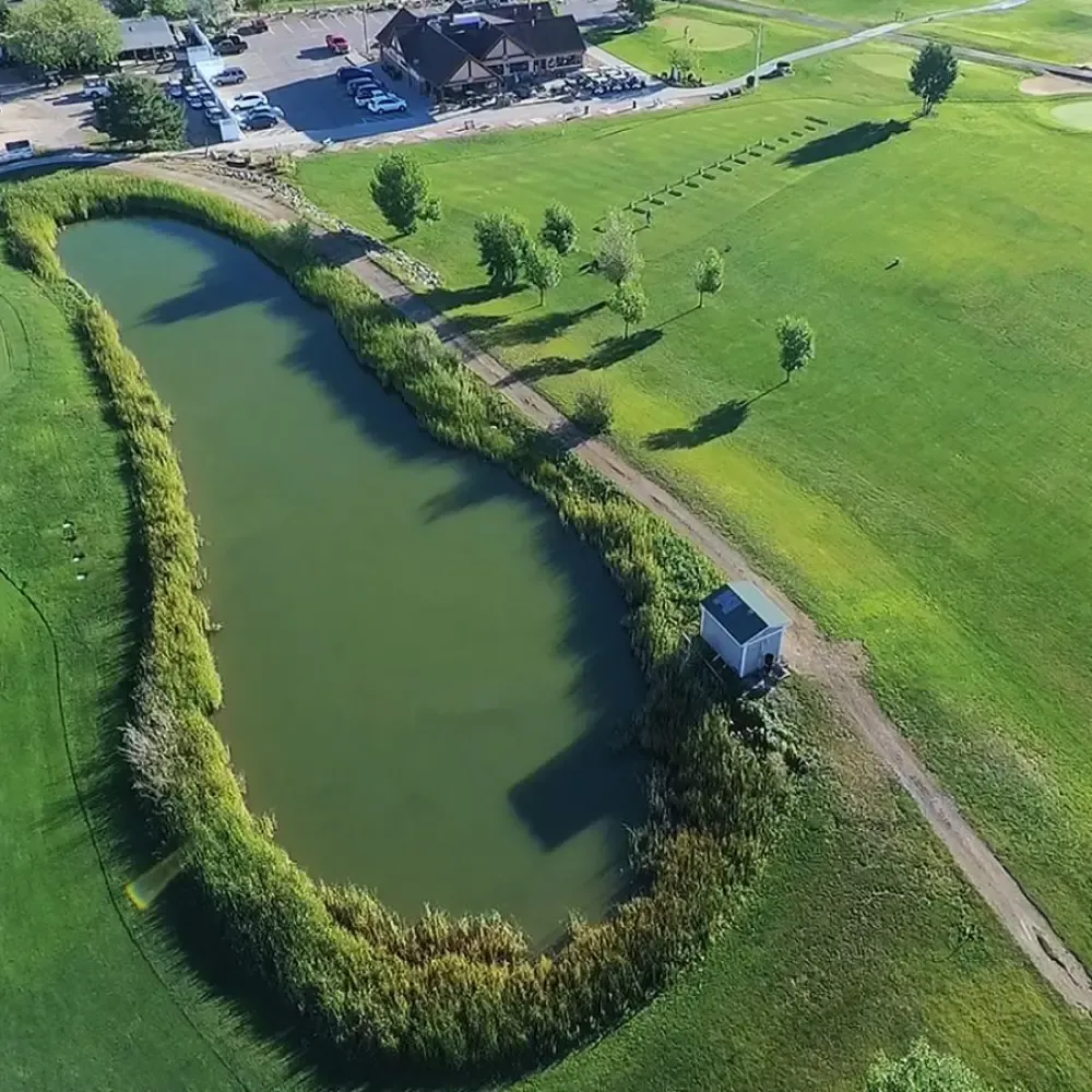 Aerial view of a pond surrounded by greenery and a pathway, with a building and parking area in the background.