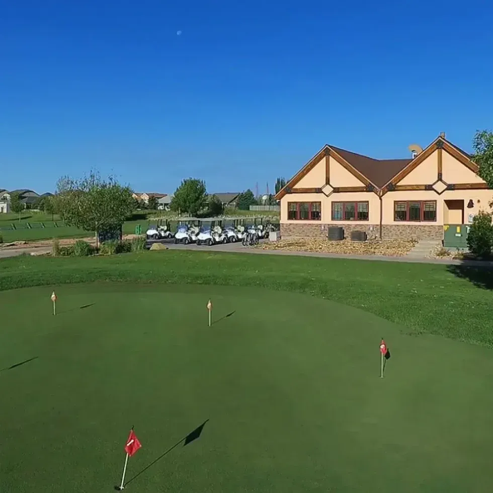 A golf course with a clubhouse in the background, featuring green grass and golf flags on the putting green.