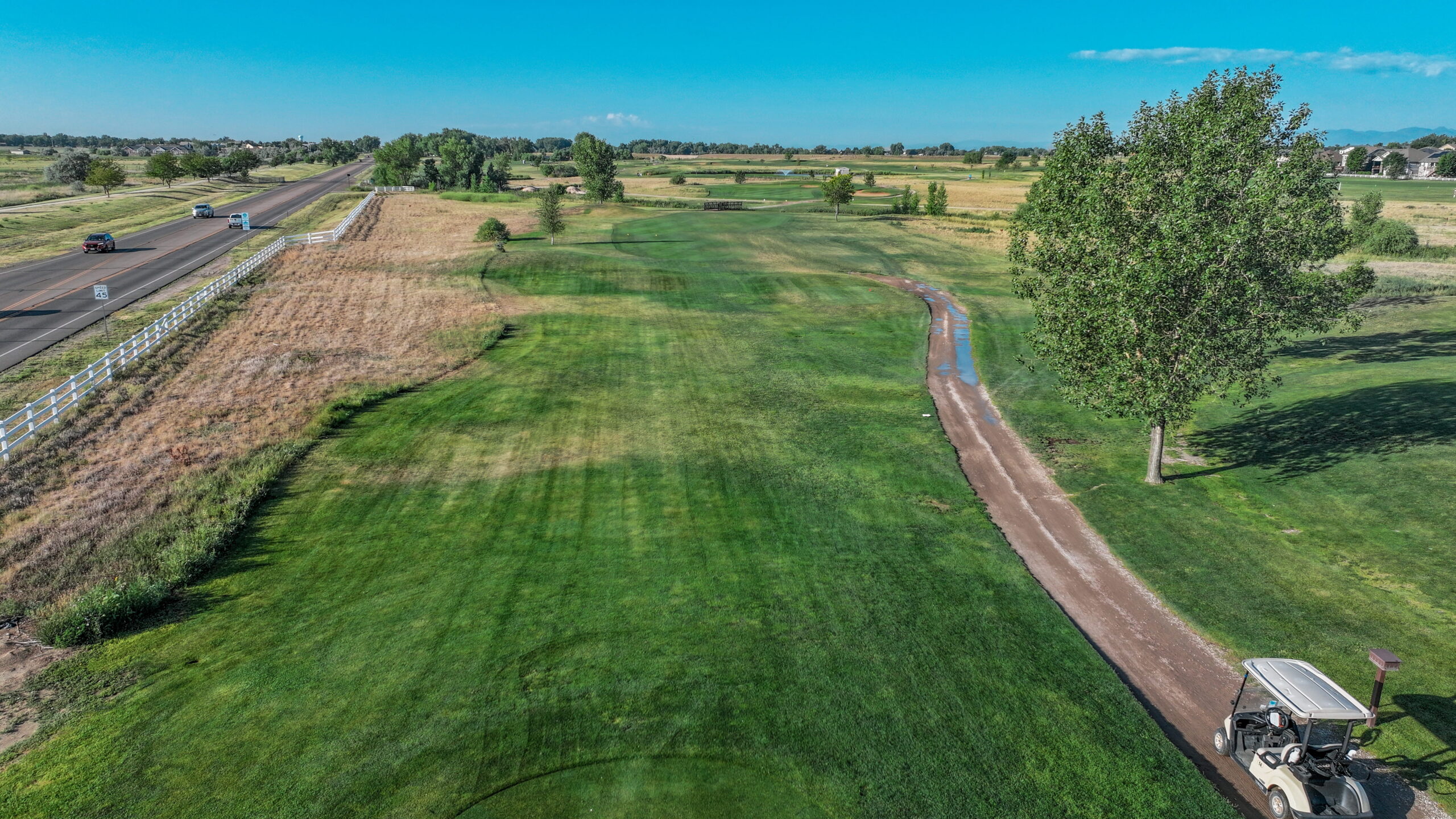 Aerial view of a golf course hole with a cart path, green grass, and a road in the background.