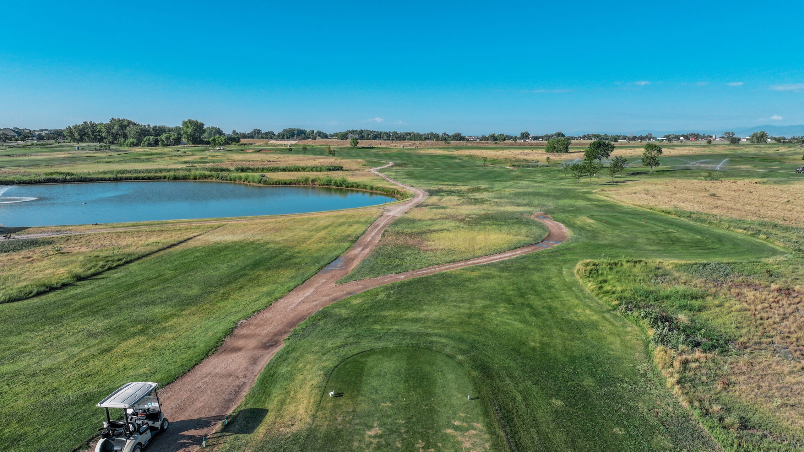 Aerial view of a golf course hole with a pond, green fairways, and winding paths under a clear blue sky.
