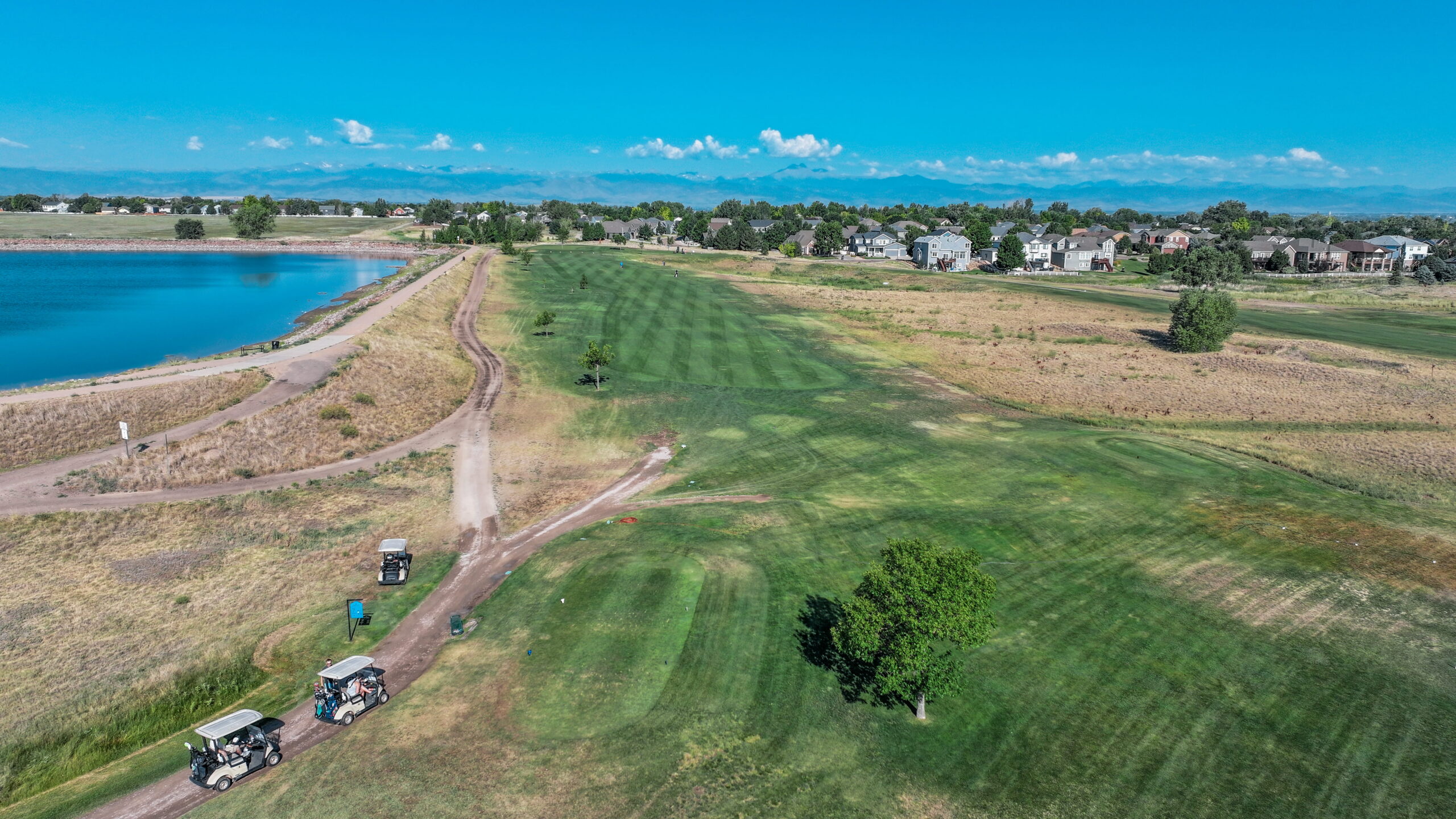 Aerial view of a golf course with golf carts on a dirt path, a lake, and residential homes in the background.