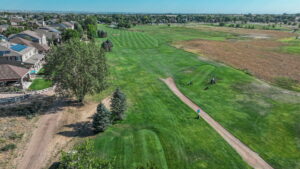 Aerial view of a golf course with green fairways, trees, and nearby houses under a clear blue sky.