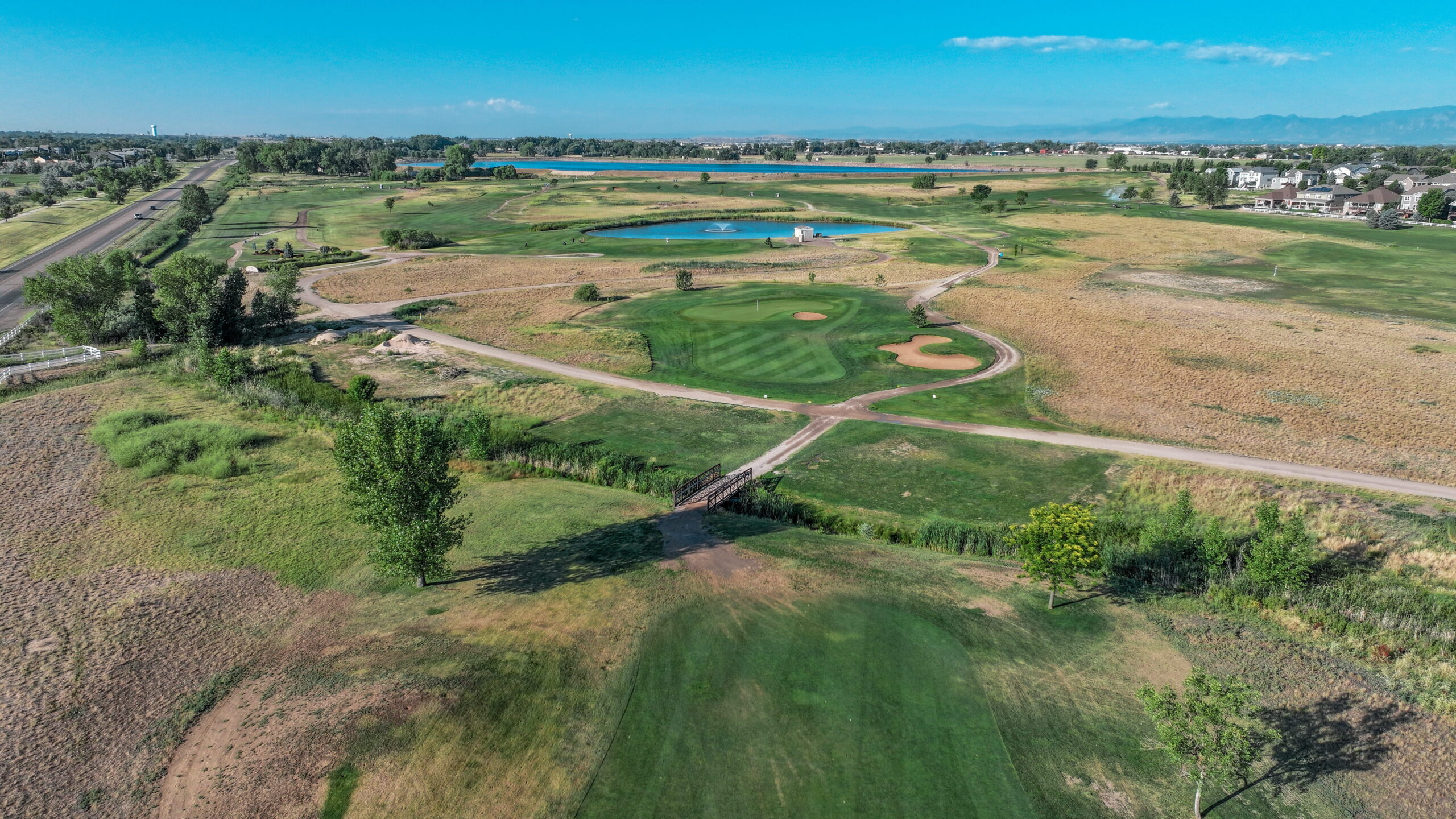 Aerial view of a golf course with green fairways, sand traps, and a pond under a clear blue sky.