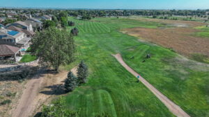 Aerial view of a golf course with green fairways, trees, and nearby houses under a clear blue sky.
